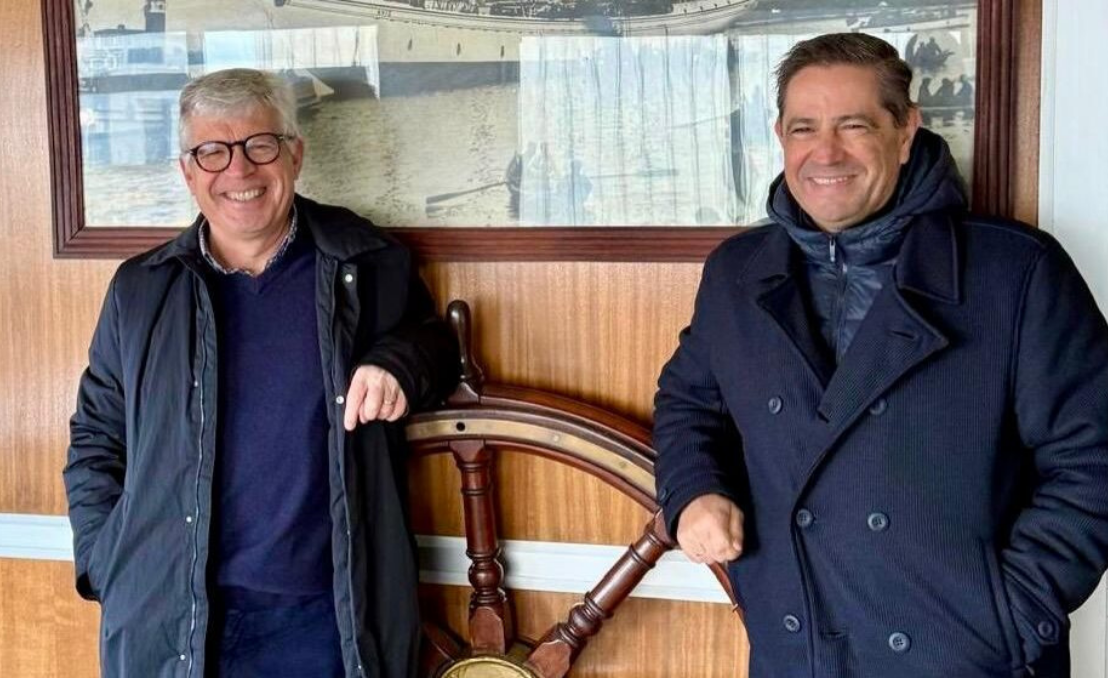 Two smiling men indoors against wood paneling, standing next to a nautical decorative element.